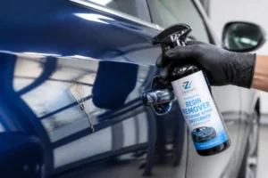 Main gantée appliquant un spray nettoyant sur une tache de sève pour nettoyer la carrosserie d'une voiture.